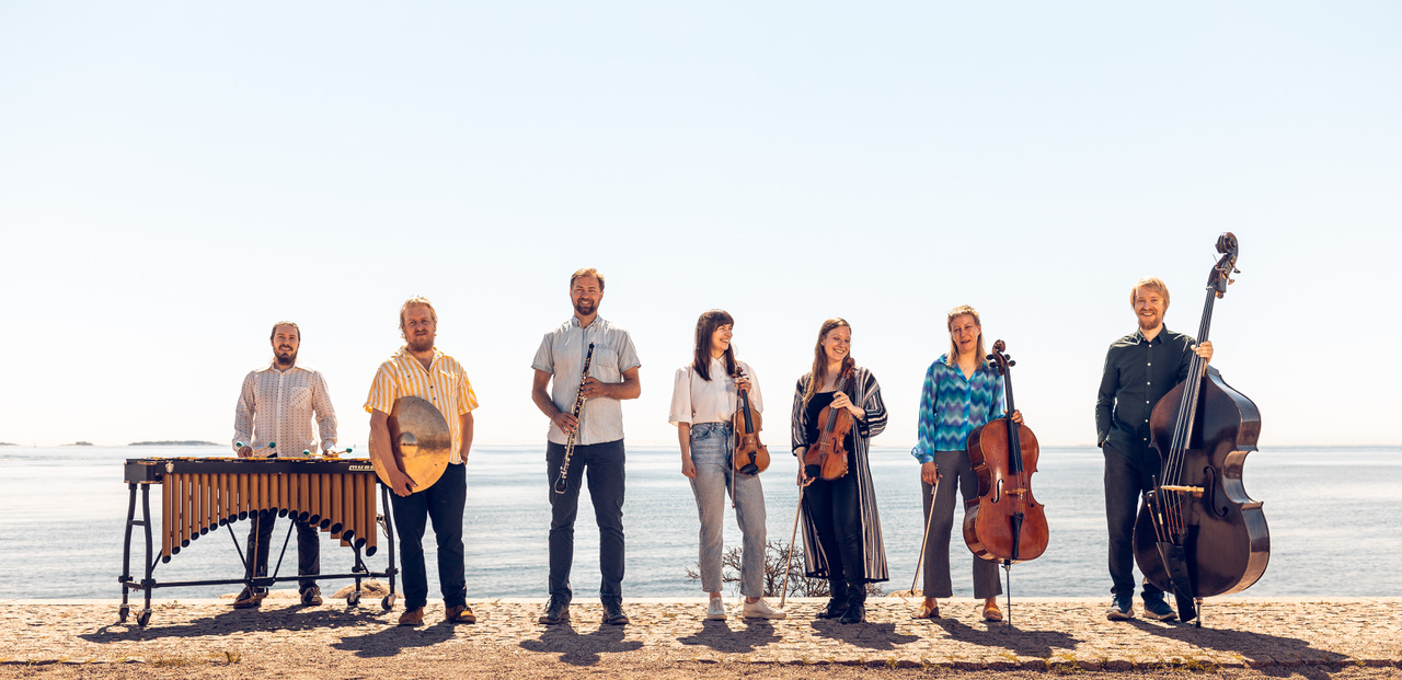 Seitsemän muusikkoa seisoo rannalla