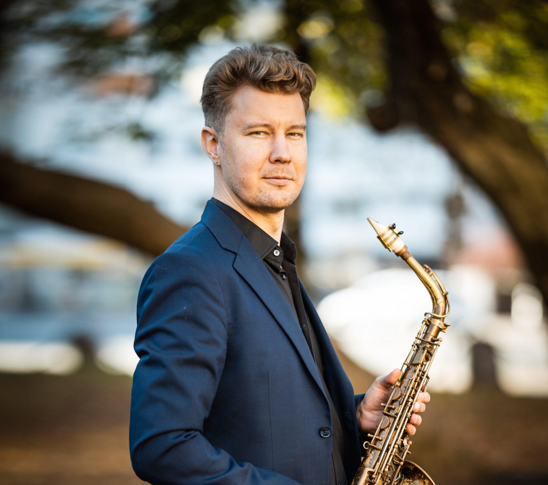 Muusikko Ville Vannemaa tummansinisessä pikkutakissa saksofoni kädessä, taustalla näkyy puustoa.